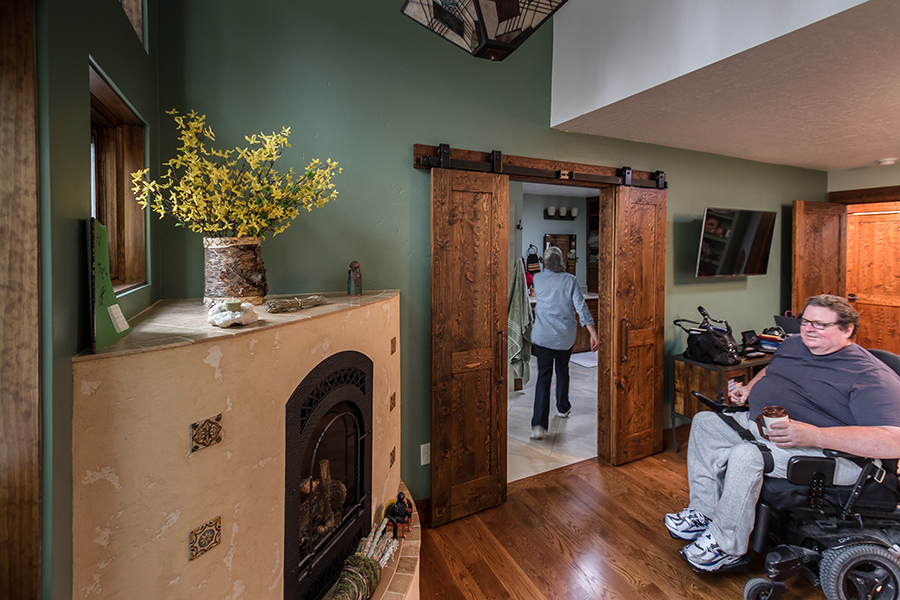 A bemused man holding coffee in a wheelchair looks at a fireplace while in a room with an open-floor plan, while his wife walks into another room through a pair of sliding barn-style doors designed to maximize usable floor space.